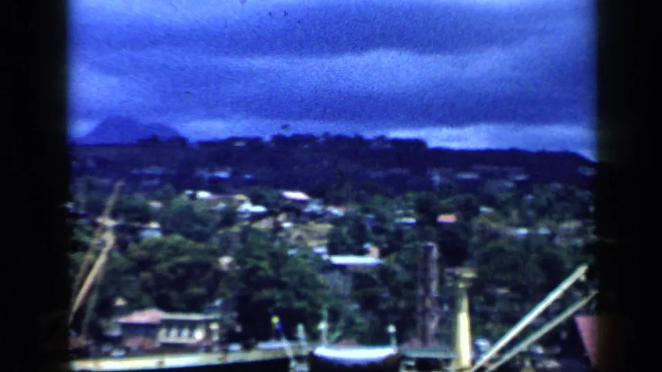 1959 Venezuela 8mm view of a mountainous urban waterfront with cranes, clustered buildings, and heavy blue cloud cover.