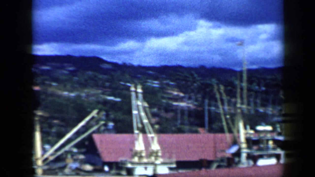 1959 Venezuela 8mm frame with a waterfront roofline, cranes, boat rigging, and a mountainous city slope in the background.