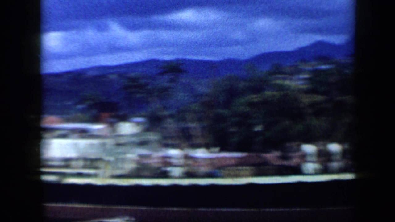 1959 Venezuela 8mm pan frame showing a blurred waterfront edge, hillside trees, distant mountains, and blue-gray overcast sky.