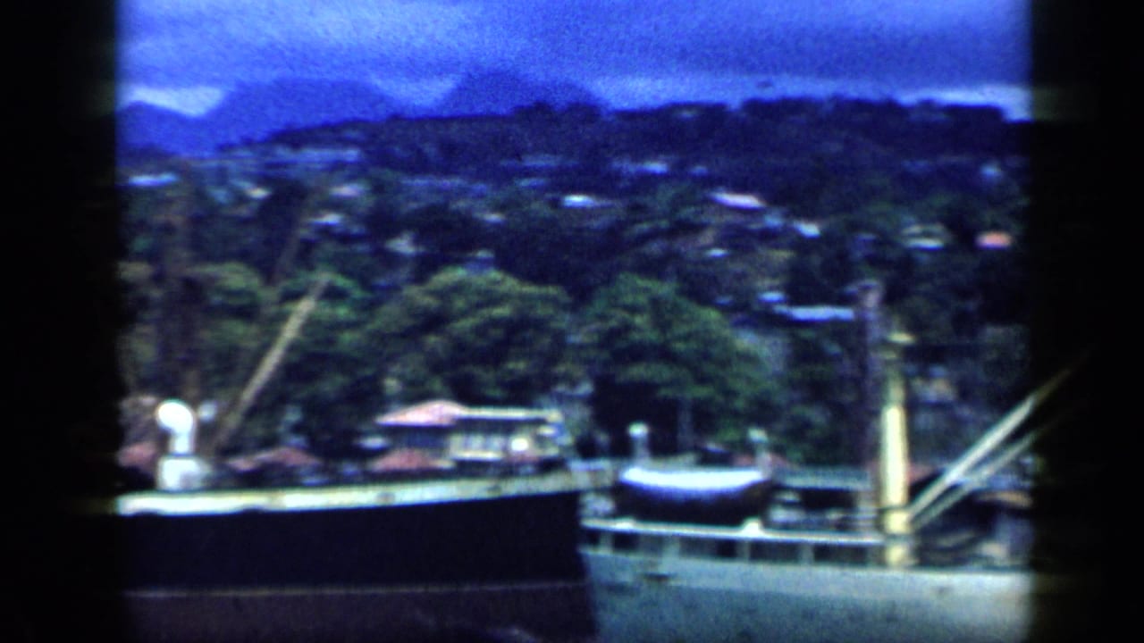 1959 Venezuela harbor view with a moored cargo ship, dockside structures, hillside houses, and dense greenery under a blue sky.