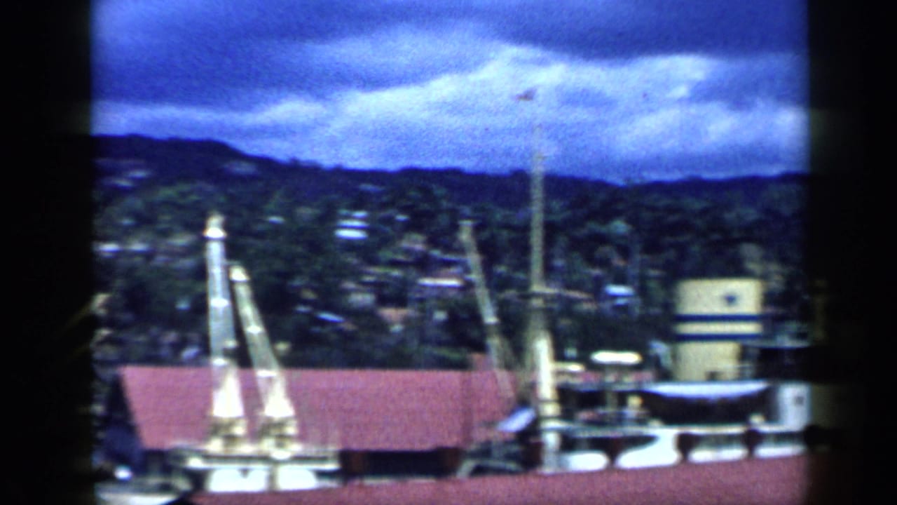 1959 Venezuela port scene with a moored vessel, dock cranes, red-roofed waterfront building, and houses rising on the hillside.