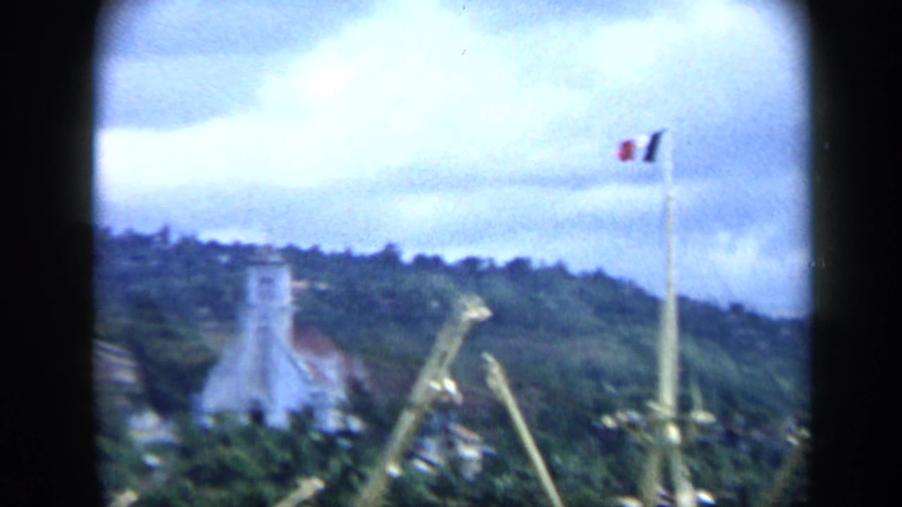 1959 Venezuela harbor view with a flagpole, tower or lighthouse-like structure, dock cranes, and green hillside beyond the waterfront.