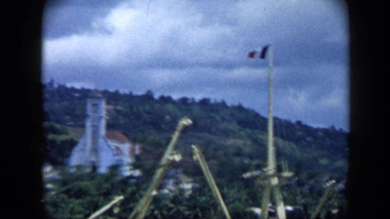 1959 Venezuela port view showing a flagpole, cranes, a tower or lighthouse-like building, wooded hillside, and overcast blue sky.