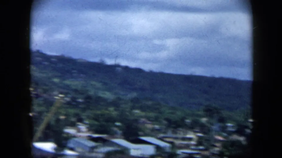 1959 Venezuela home-movie frame showing a lush hillside with scattered buildings beneath a cloudy sky, seen from a moving viewpoint.