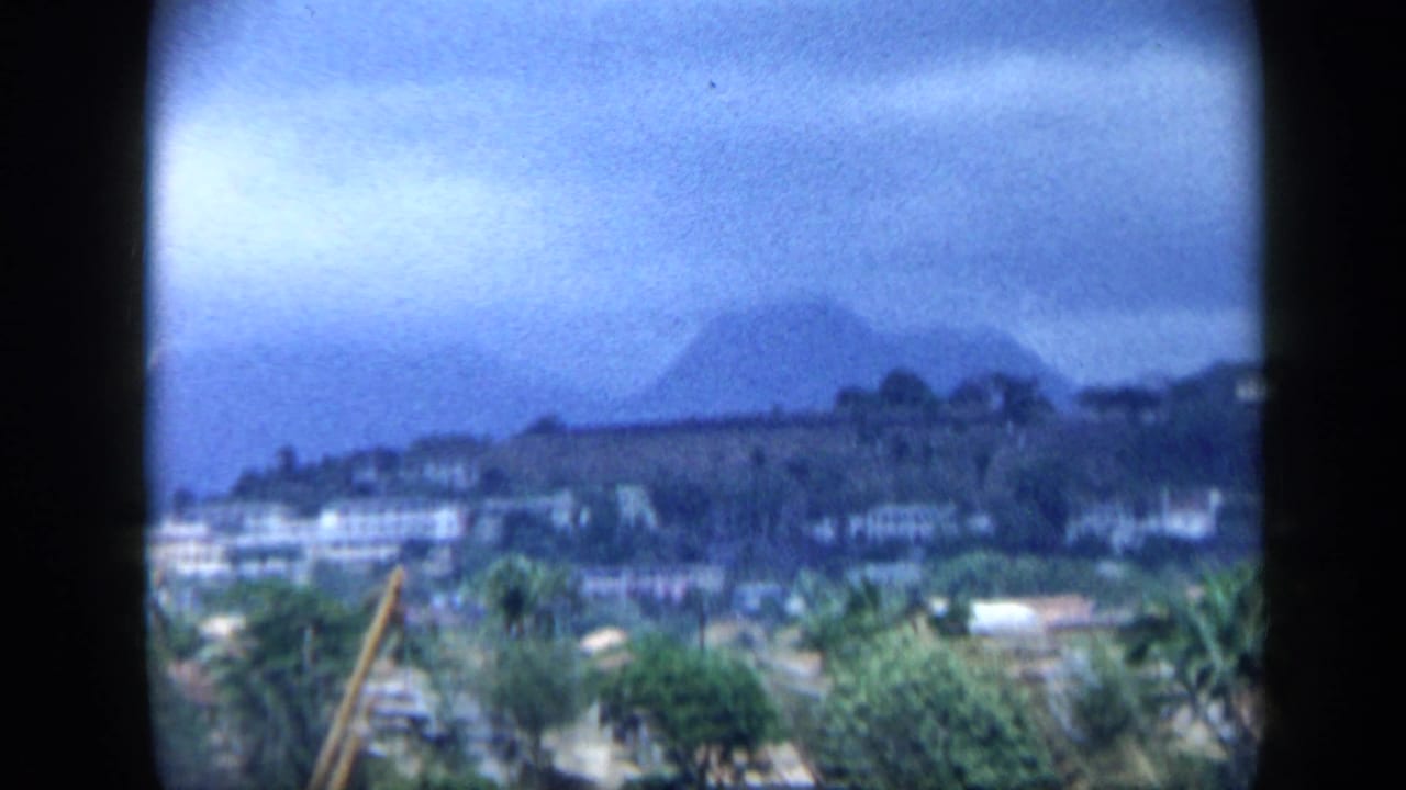 1959 Venezuela home-movie frame showing a distant waterfront settlement, palm trees, and dark mountain forms through soft haze.
