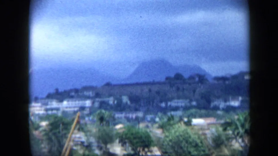 1959 Venezuela home-movie frame showing a distant waterfront settlement, palm trees, and dark mountain forms through soft haze.