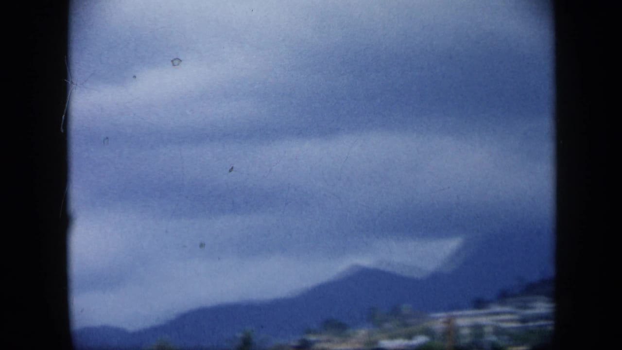 1959 Venezuela travel frame showing a dark mountain ridge, low clouds, and a small strip of buildings near the lower edge.