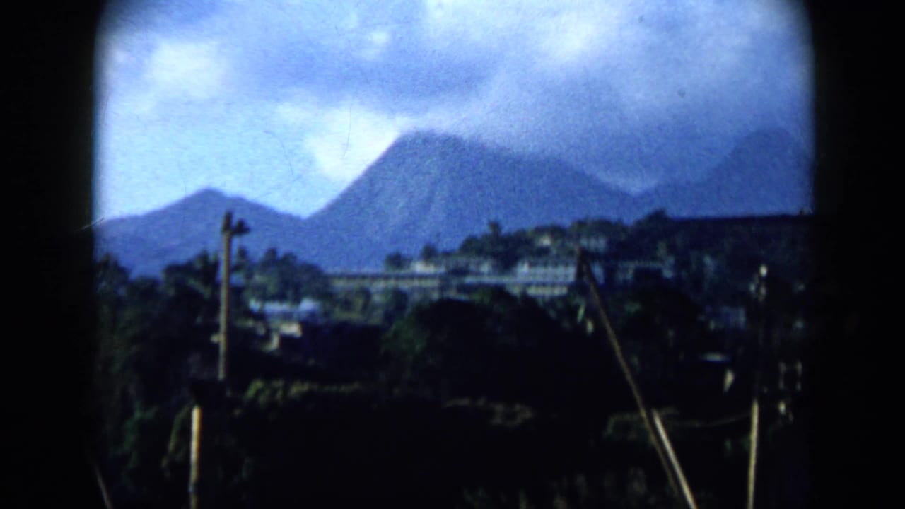 1959 Venezuela 8mm view of a mountain-backed settlement with hillside buildings, vegetation, and utility poles beneath a cloudy sky.
