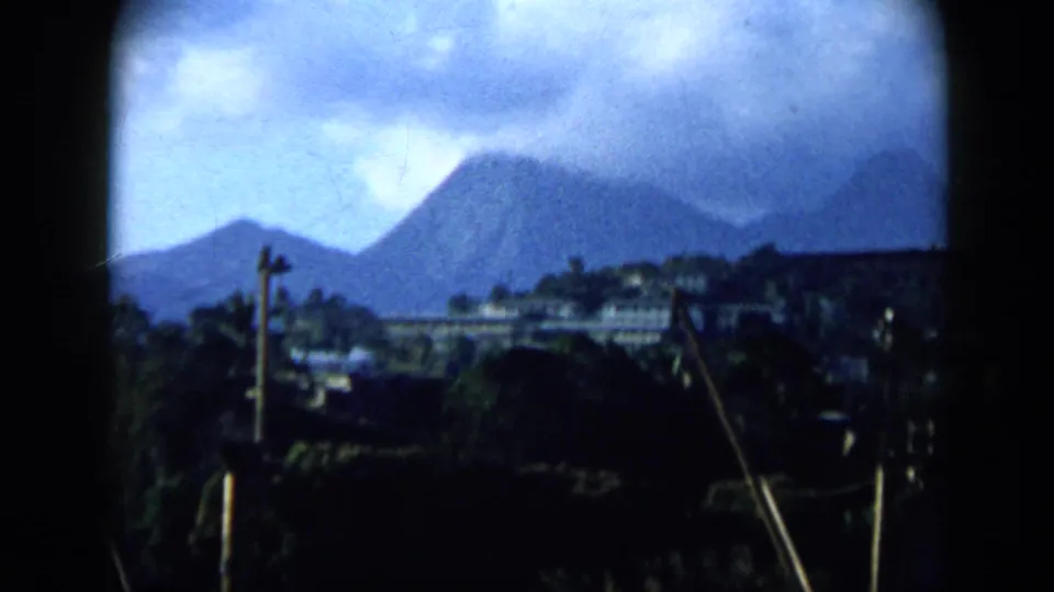 1959 Venezuela 8mm view of a mountain-backed settlement with hillside buildings, vegetation, and utility poles beneath a cloudy sky.
