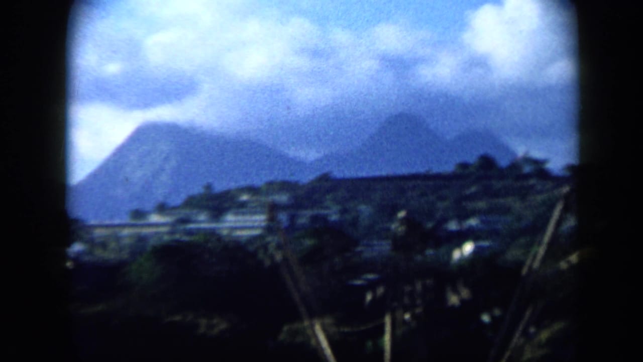 1959 Venezuela 8mm frame showing a cloudy mountain backdrop, low hillside buildings, palms, and diagonal utility lines or poles.