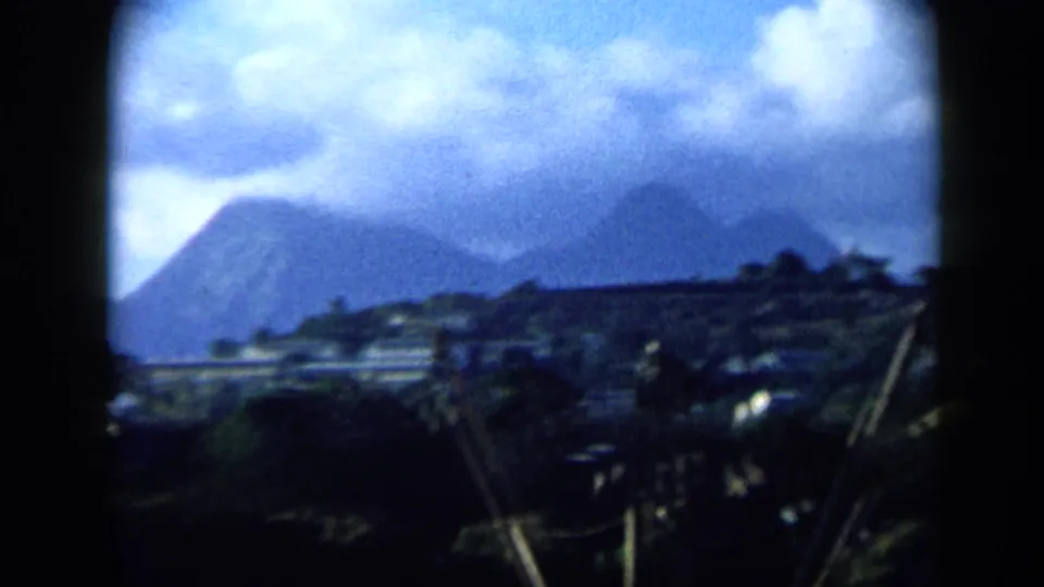 1959 Venezuela 8mm frame showing a cloudy mountain backdrop, low hillside buildings, palms, and diagonal utility lines or poles.