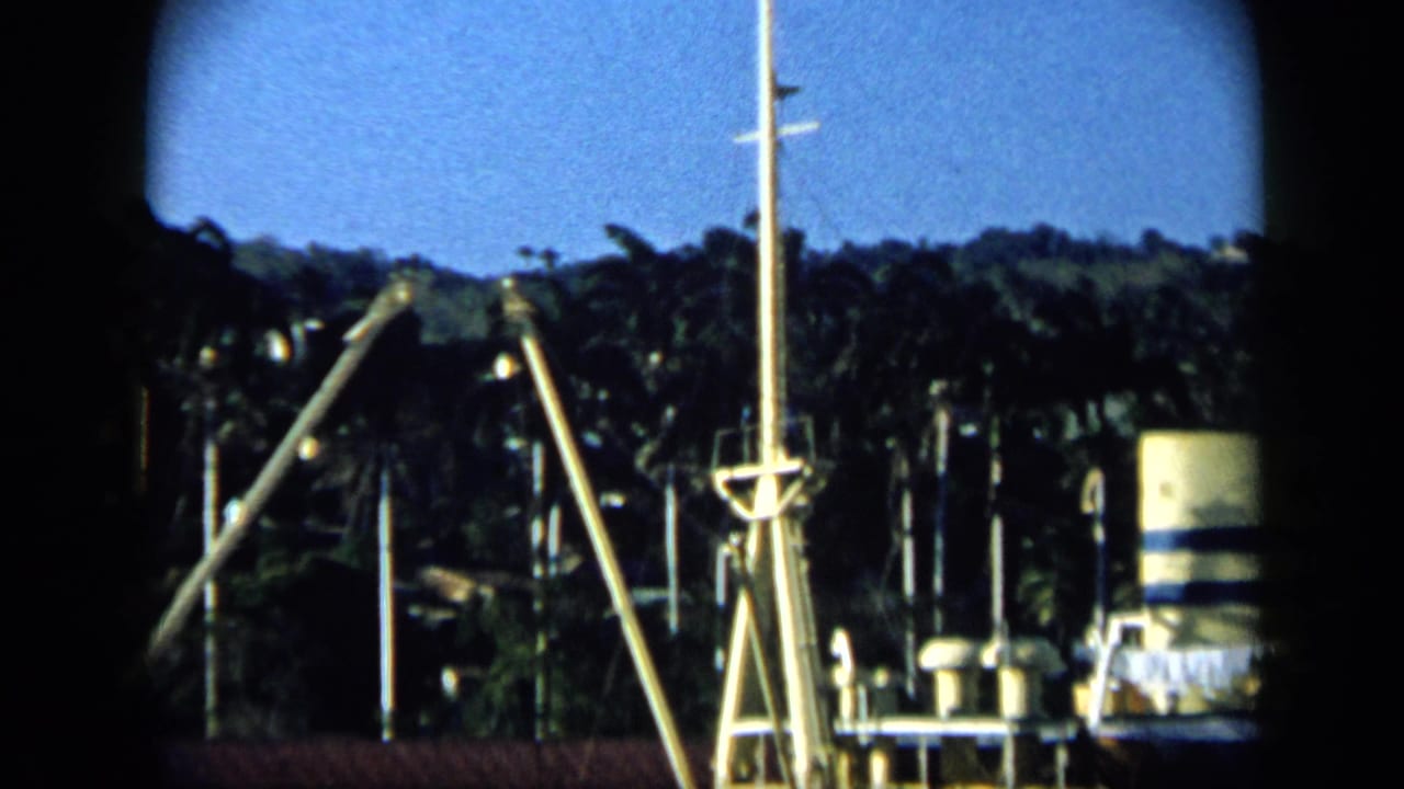 1959 Venezuela 8mm frame showing boat rigging before a green shoreline, with a cylindrical shore tank and blue sky.