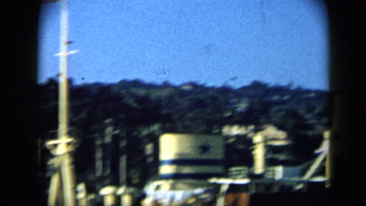 1959 Venezuela boat-view frame with a tall mast, rigging, blurred waterfront tank, and wooded hillside.