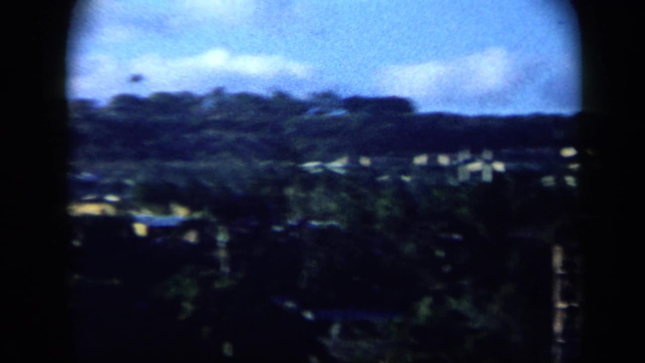 1959 Venezuela shoreline frame showing a soft, handheld view of hillside, scattered shore buildings, and blue sky.