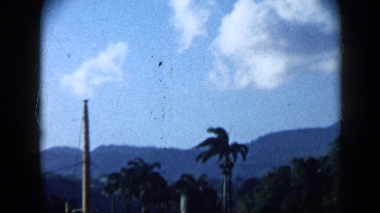 1959 Venezuela frame with palms, distant mountain ridges, bright clouds, and a tall vertical industrial or utility element.