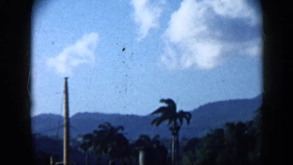 1959 Venezuela frame with palms, distant mountain ridges, bright clouds, and a tall vertical industrial or utility element.