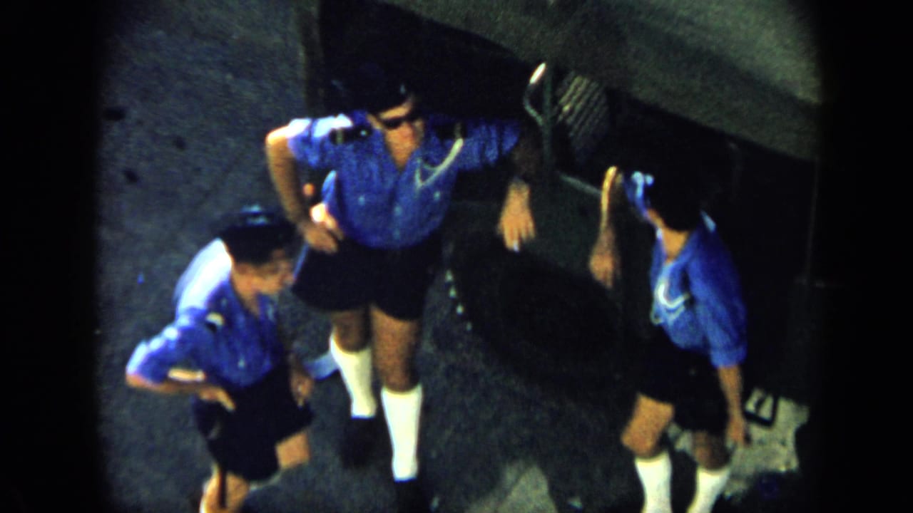 Overhead 1959 Venezuela home-movie view of three men in matching blue shirts and dark shorts gathered on a paved surface.