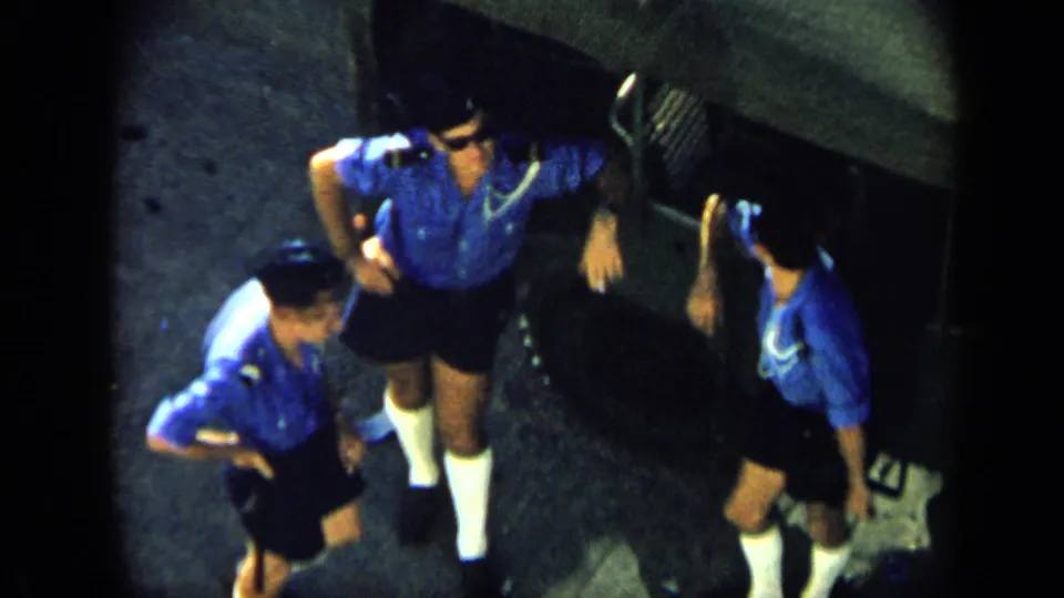 Overhead 1959 Venezuela home-movie view of three men in matching blue shirts and dark shorts gathered on a paved surface.