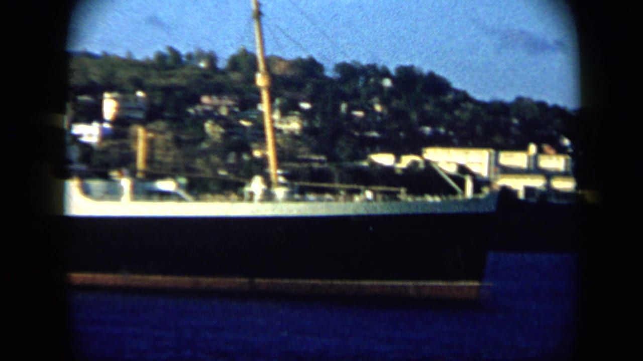 Handheld 8mm frame of a Venezuelan harbor waterfront, hillside buildings, trees, and a mast during the pan.
