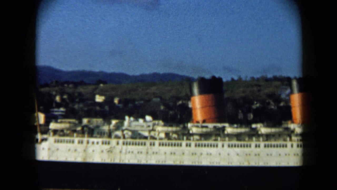 Large white passenger ship docked or near shore in Venezuela, with red-and-black funnels and hillside buildings behind it.