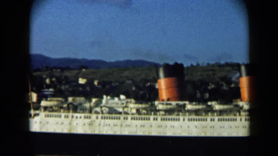 Large white passenger ship docked or near shore in Venezuela, with red-and-black funnels and hillside buildings behind it.