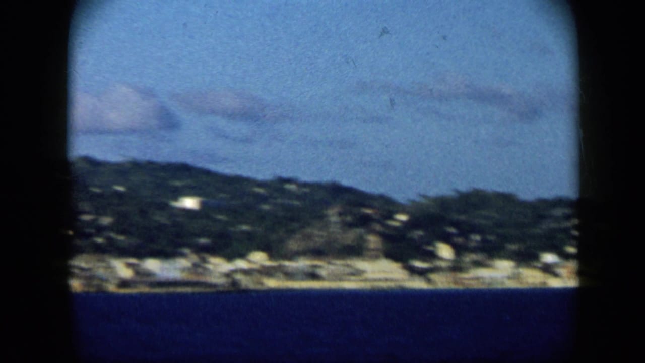 Venezuela coastal harbor view from the 1959 8mm clip, with blue water, green hillside, and small shoreline buildings.