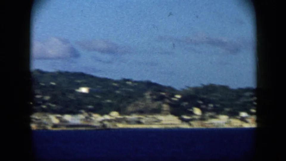 Venezuela coastal harbor view from the 1959 8mm clip, with blue water, green hillside, and small shoreline buildings.