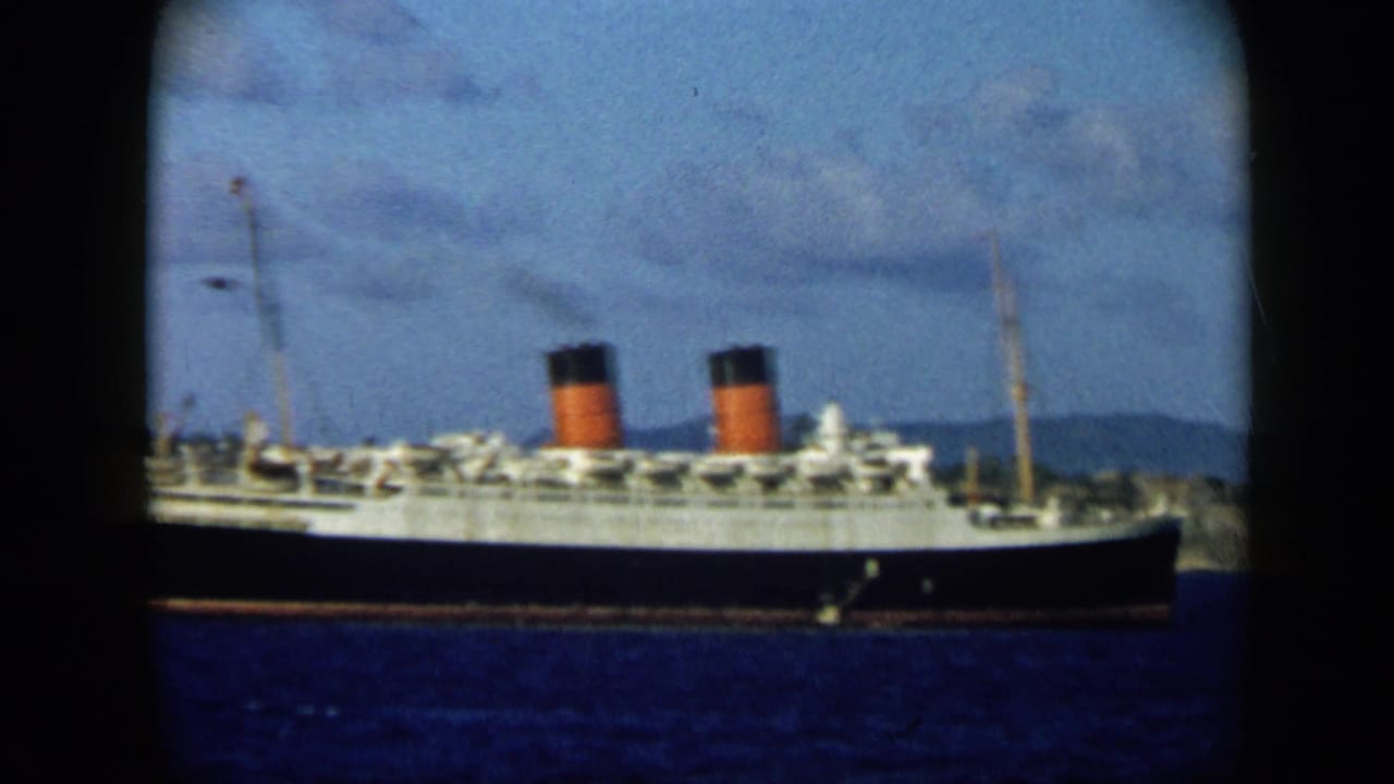 The ocean liner remains centered in the frame, showing twin red-and-black funnels, white decks, blue water, and distant coastal hills.