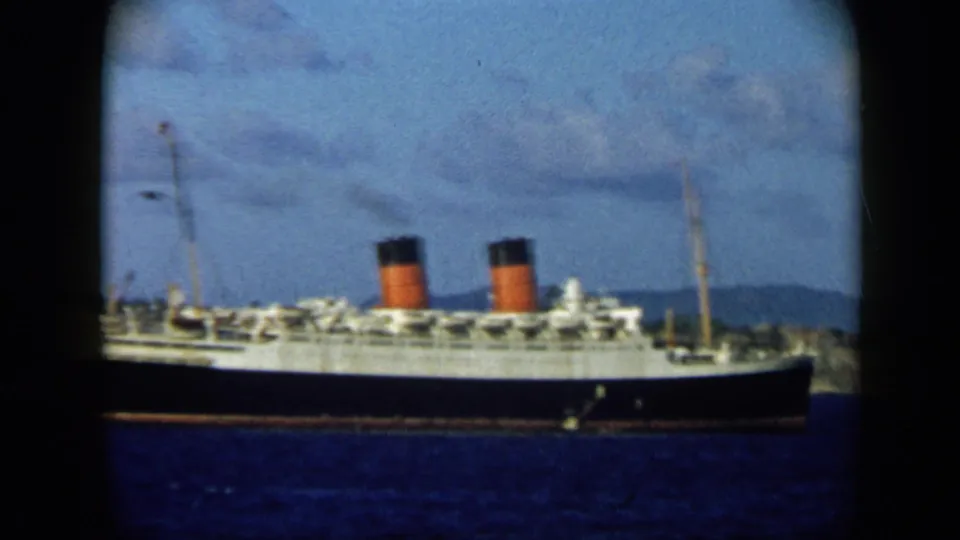 The ocean liner remains centered in the frame, showing twin red-and-black funnels, white decks, blue water, and distant coastal hills.