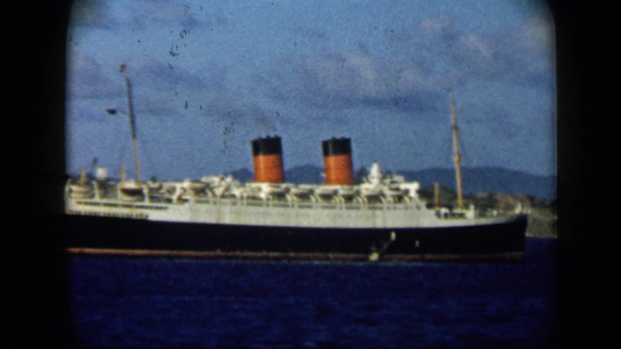 A closer 8mm view of the liner emphasizes its dark hull, white upper decks, twin funnels, blue water, and distant shoreline.