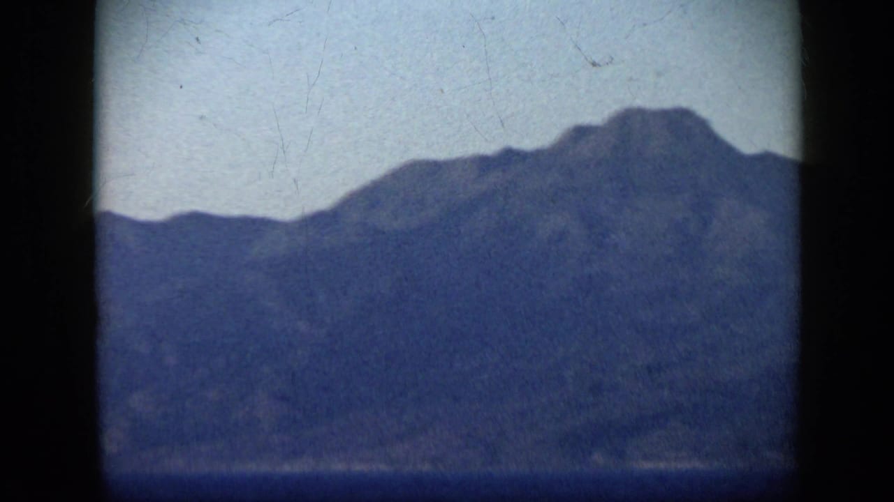 1959 Venezuela home-movie frame showing a hazy mountain profile across blue water, with the ship no longer visible.