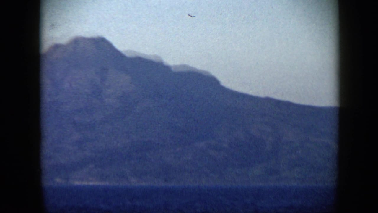 Large mountain mass above dark water with a pale sky and rounded film-frame edges.