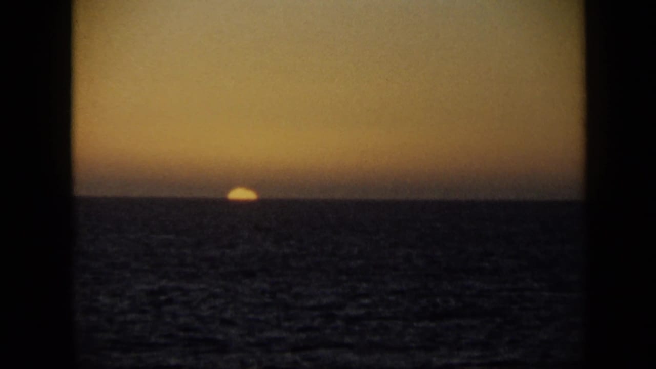 Small sun near the horizon over dark water with an amber sky and black frame edges.