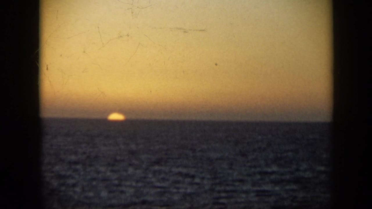 1959 Venezuela 8mm frame showing the sun nearly meeting the sea horizon beneath an amber sky and visible film scratches.