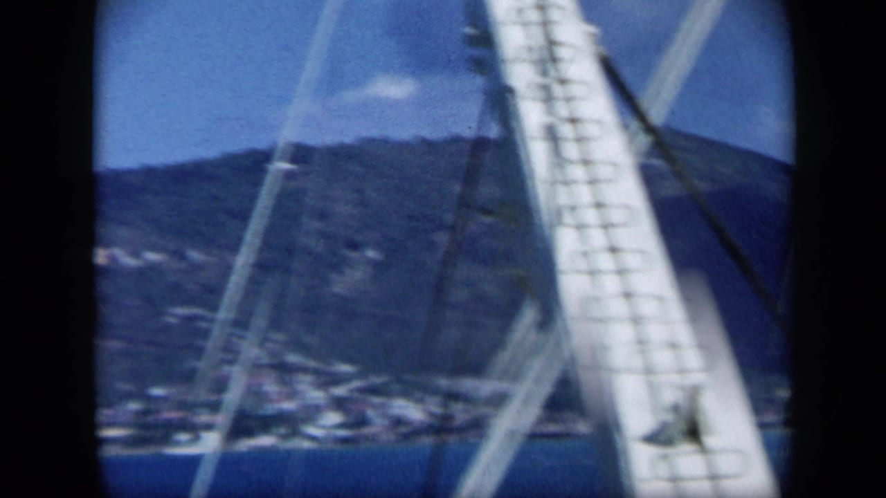 Boat rigging crosses a distant green hillside and shoreline under blue sky.