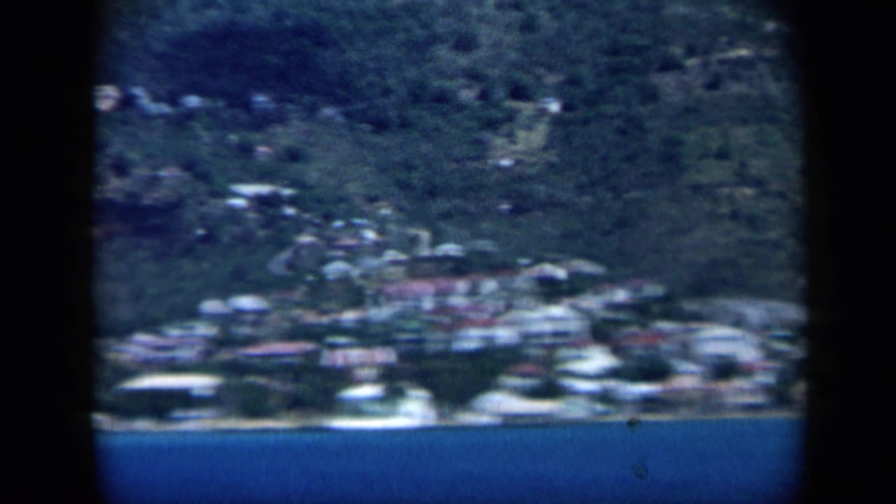 1959 Venezuela frame showing hillside buildings and shoreline greenery across blue coastal water.