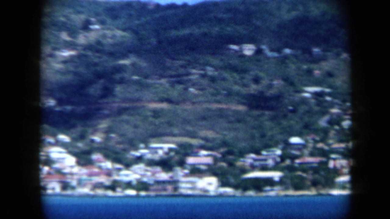 1959 Venezuela 8mm view of a seaside hillside town across blue water, with vegetation and pale buildings visible through dark frame borders.
