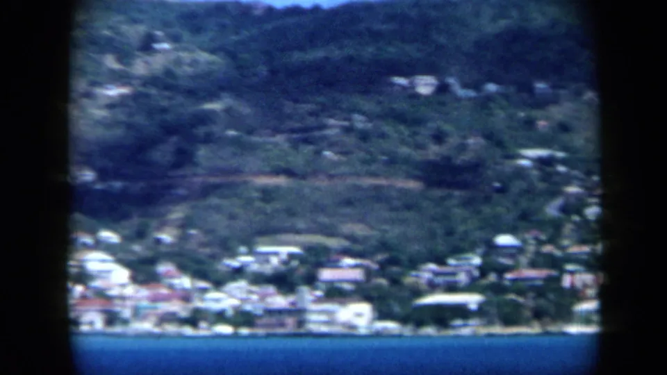 1959 Venezuela 8mm view of a seaside hillside town across blue water, with vegetation and pale buildings visible through dark frame borders.