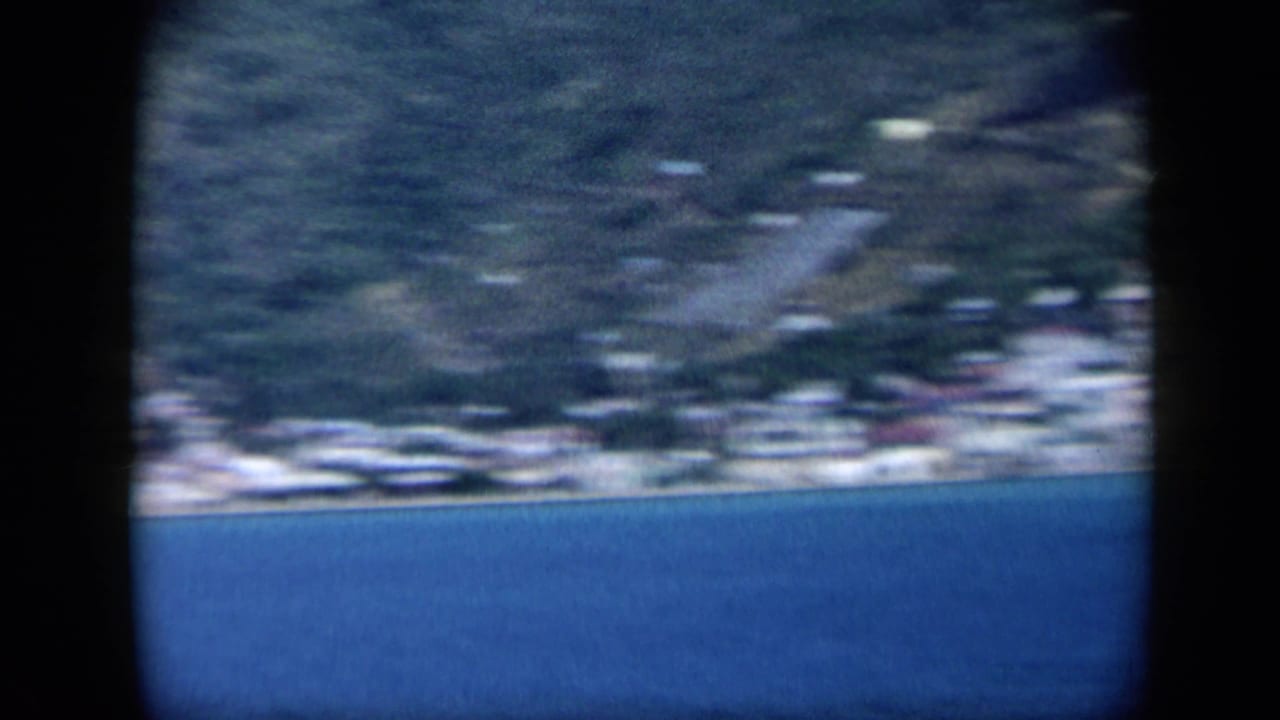 Soft 8mm view of a Venezuela coastal settlement, with blue water in the foreground and hillside buildings blurred by motion or focus.