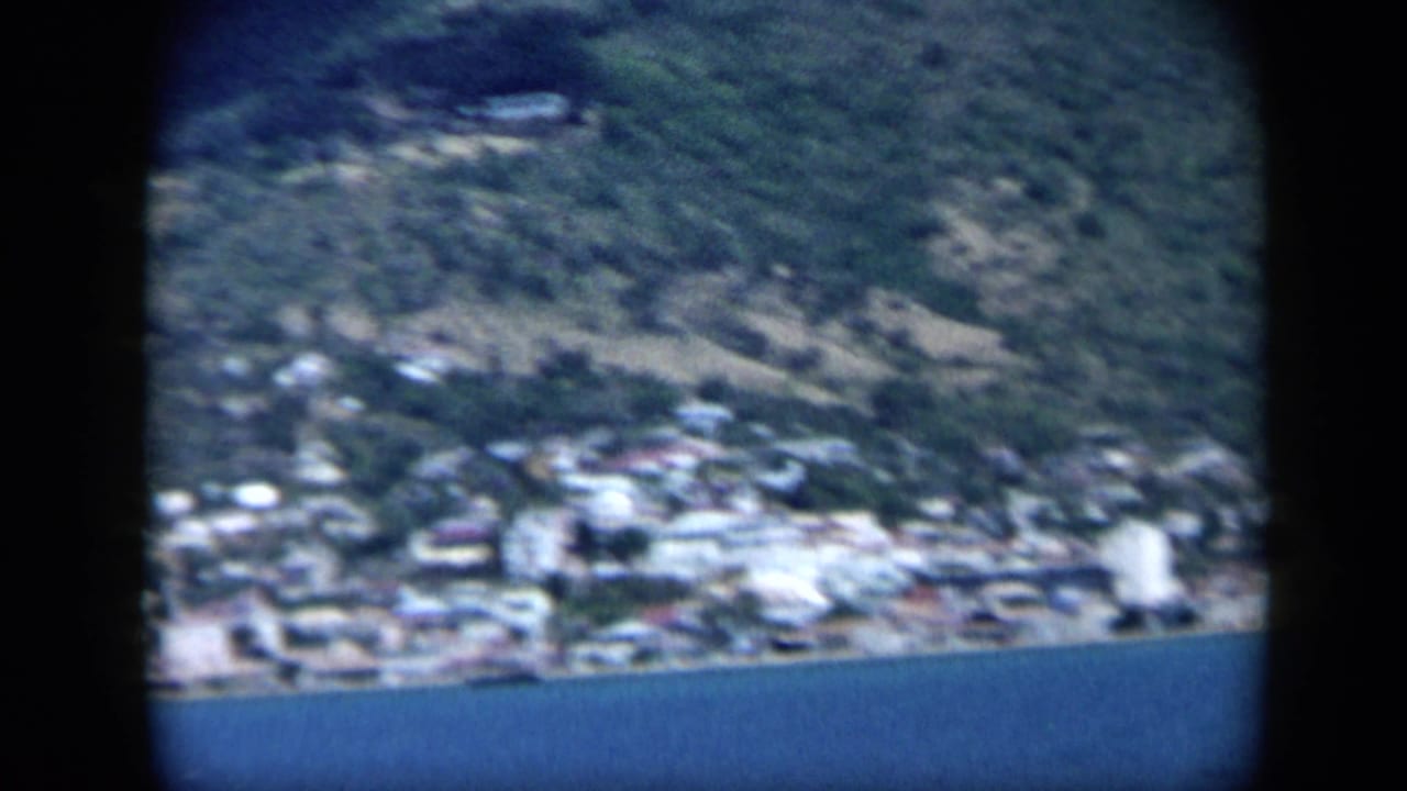 1959 Venezuela 8mm frame of a compact seaside settlement beneath a steep green hillside, with blue bay water along the lower edge.