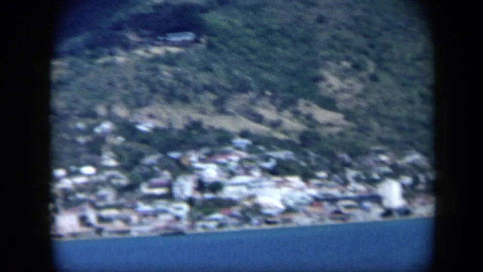 1959 Venezuela 8mm frame of a compact seaside settlement beneath a steep green hillside, with blue bay water along the lower edge.