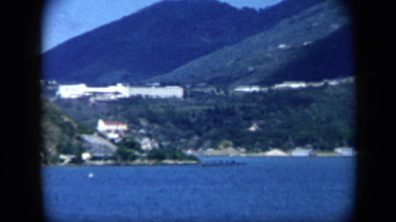 1959 8mm view across a Venezuelan bay toward white waterfront and hillside buildings beneath green slopes.