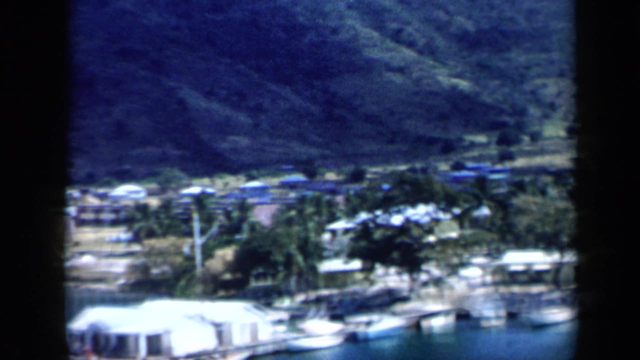 The pan continues over a Venezuelan coastal inlet, showing small boats, shoreline structures, palms, utility poles, and a shaded mountain backdrop.