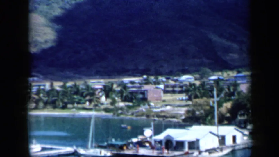 A closer 8mm frame shows a marina edge in Venezuela with white waterside structures, docked boats, palms, a road, and the steep hillside beyond.