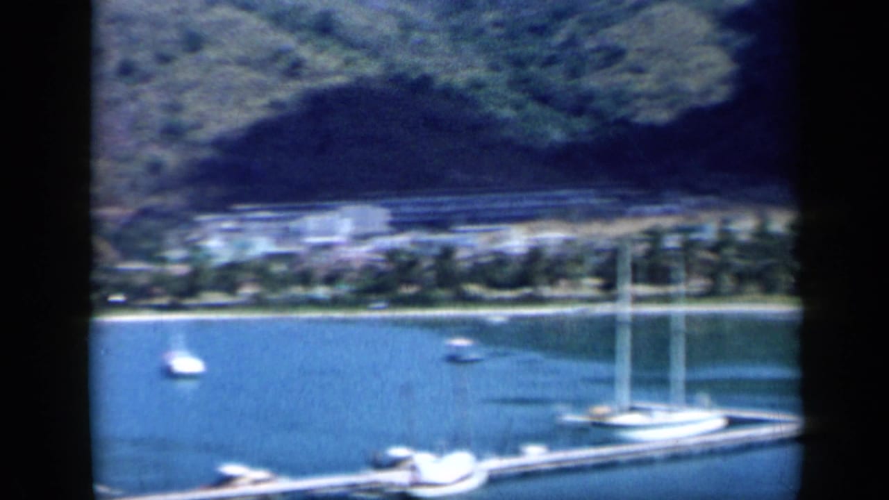 The final frame opens onto calm blue water with a sailboat, smaller boats, palm-lined shore, low buildings, and a dark green mountain behind.