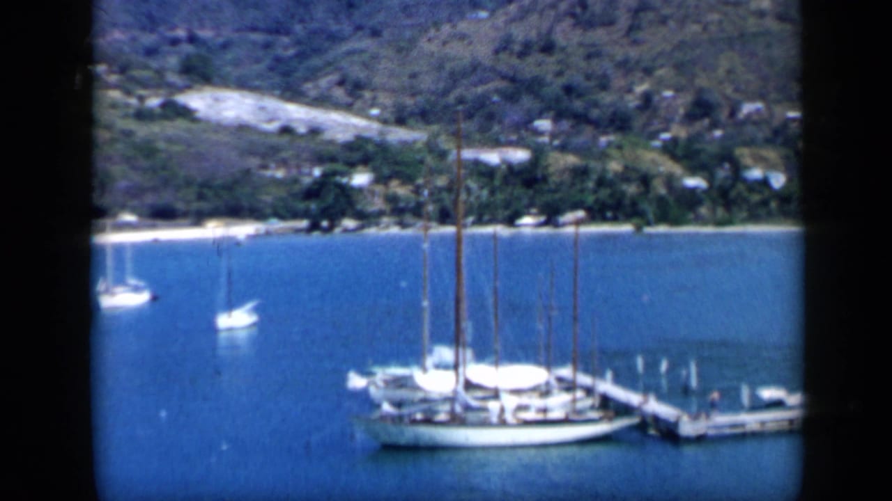 1959 Venezuela home-movie frame showing anchored sailboats, calm harbor water, shoreline trees, and green mountains beyond.
