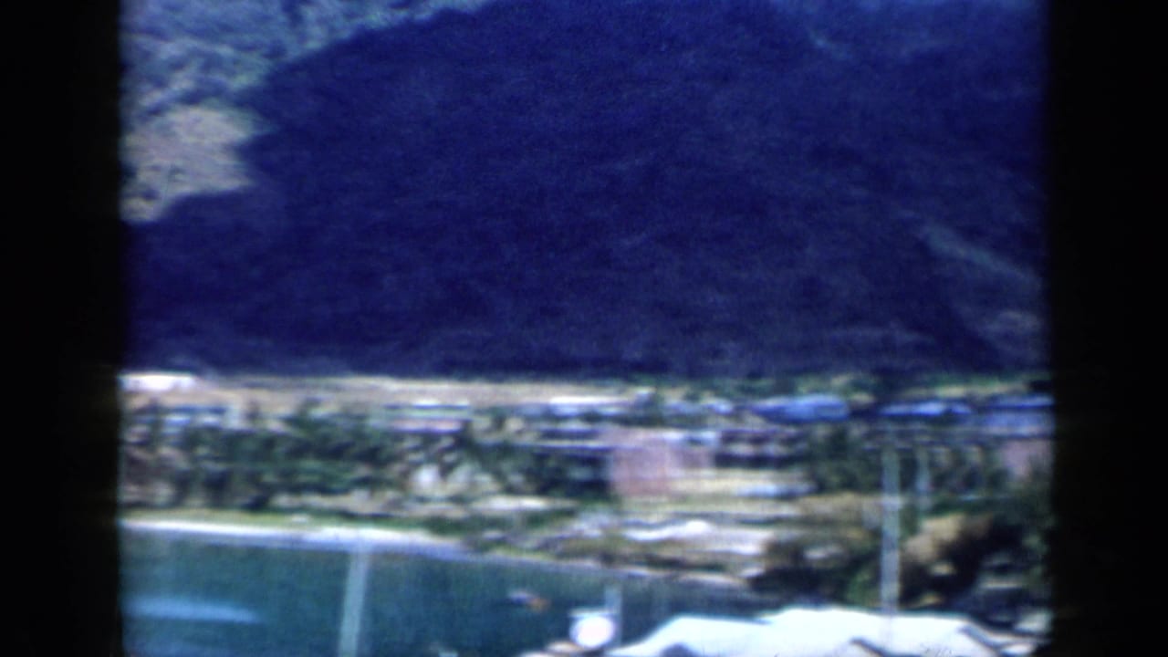1959 Venezuela frame looking across a tropical bay toward a shoreline settlement backed by green mountains.