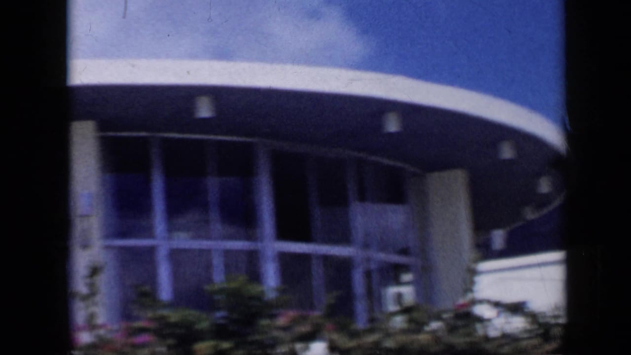 1959 Venezuela 8mm frame showing a curved modernist entrance canopy, vertical columns, glass panels, and low greenery.