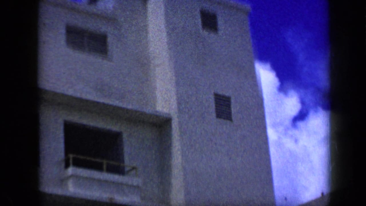 1959 Venezuela 8mm frame showing a tall white building corner with small windows, balcony recesses, and bright blue sky.