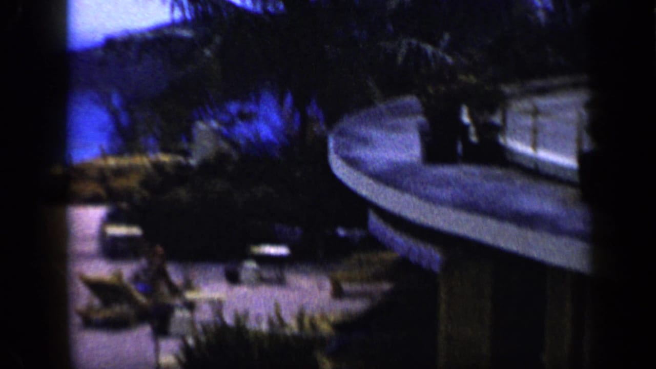 Grainy 1959 Venezuela resort view with lounge chairs, palm vegetation, terrace paving, and part of a hotel structure.
